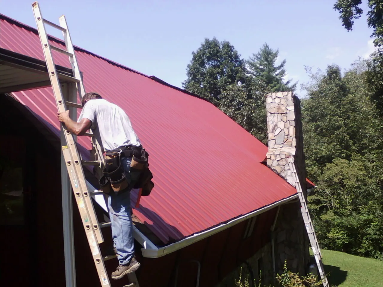 Roofer on a ladder beside a red metal roof and stone chimney — Soffit & Fascia Repair by Roofing Brothers in Register